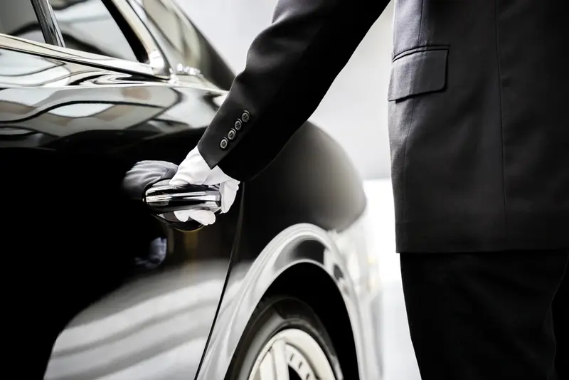 A professional driver holds the handle of the rear door of a luxurious black sedan to open it for the customer. This symbolize Houston Private Transportation.